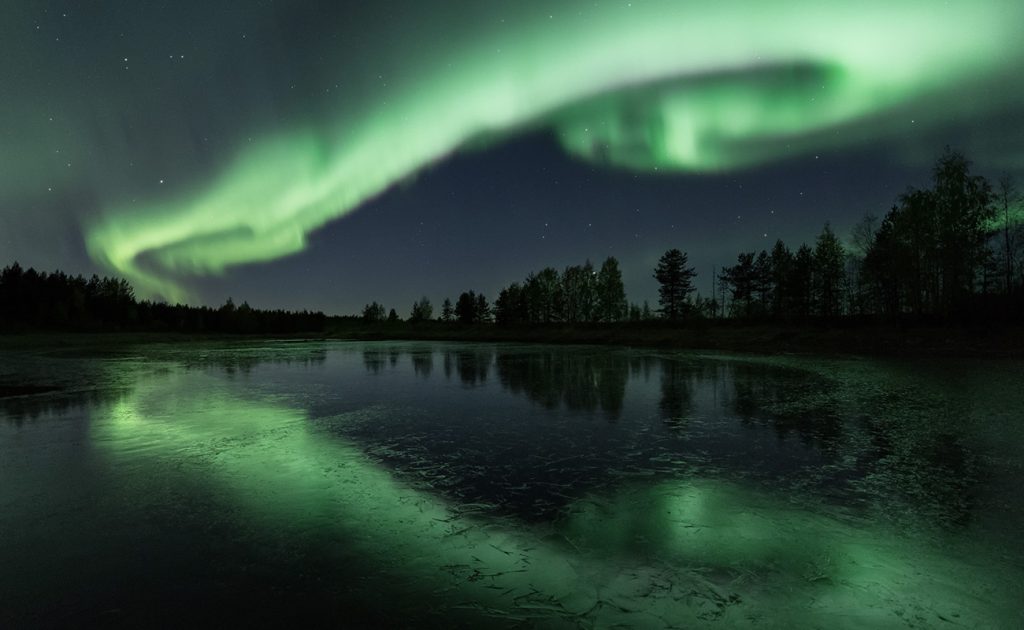 Ulkomaisema yöllä. Vihreänä loistavat revontulet heijastuvat taivaalta jäätyneen lammen pintaan.