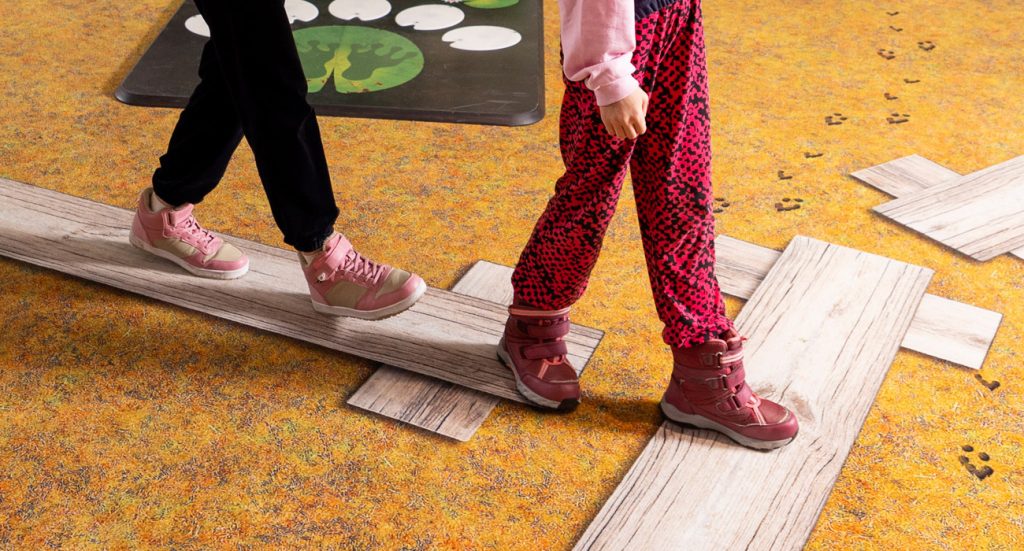 Close-up of feet. Two children are walking along duckboards. The duckboards are not real but printed on wall-to-wall carpet.