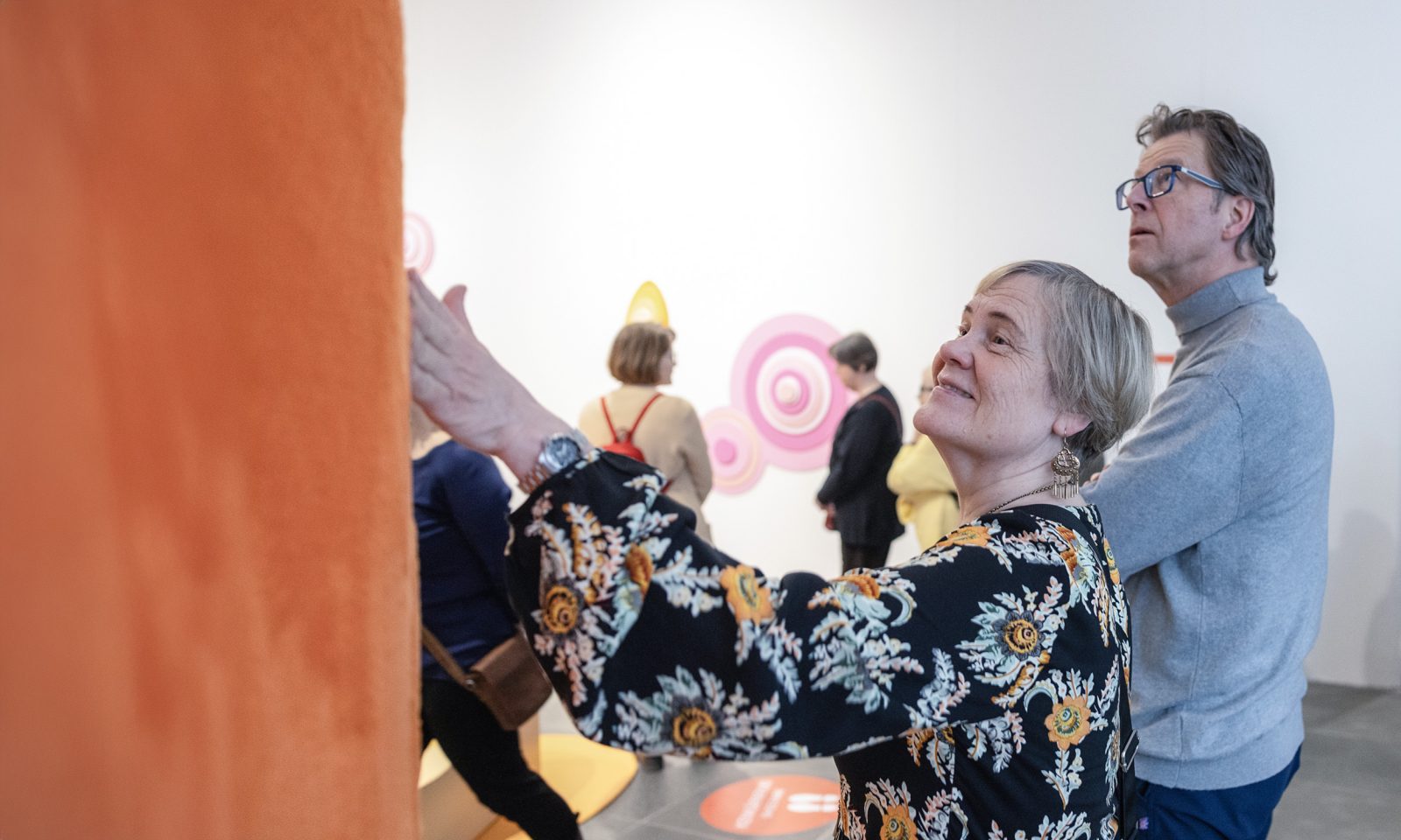 Seniori-ikäinen henkilö koskettaa karvaisen taideteoksen pintaa hymyillen.