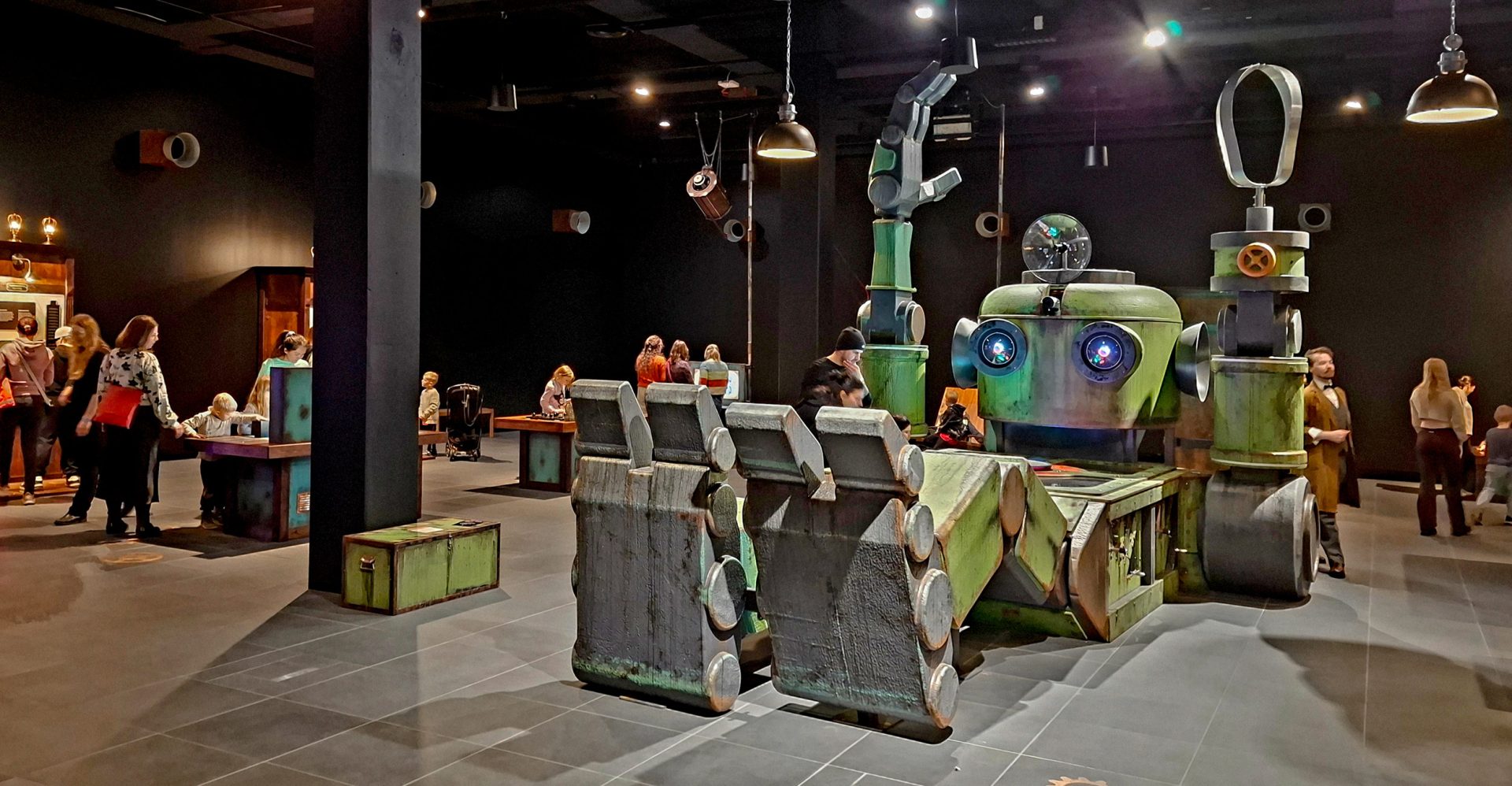 The dimly lit exhibition hall of Tietomaa’s MASIINA exhibition, featuring a large greenish-grey robot at its centre. The robot has glowing blue eyes, enormous mechanical hands and raised arms. In the background, children and adults explore the exhibition’s interactive exhibits.